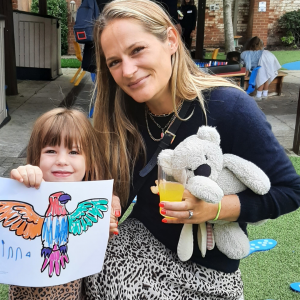 A mother and her child with a teddy bear in her hand and her daughter holding up a drawing of a parrot coloured in