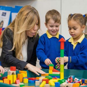 Woman and two children building colorful blocks at a teal table in a classroom with a supervising adult nearby.