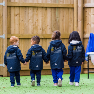 Children in navy Holy Cross jackets stand with their backs to the camera in front of a wooden fence.