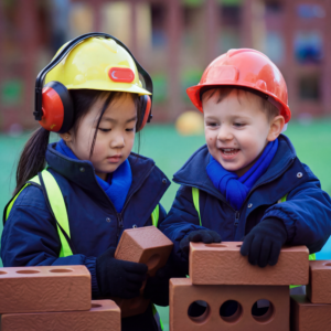 Two children in construction gear (yellow and orange helmets, reflective vests) stack brown brick-shaped blocks together.